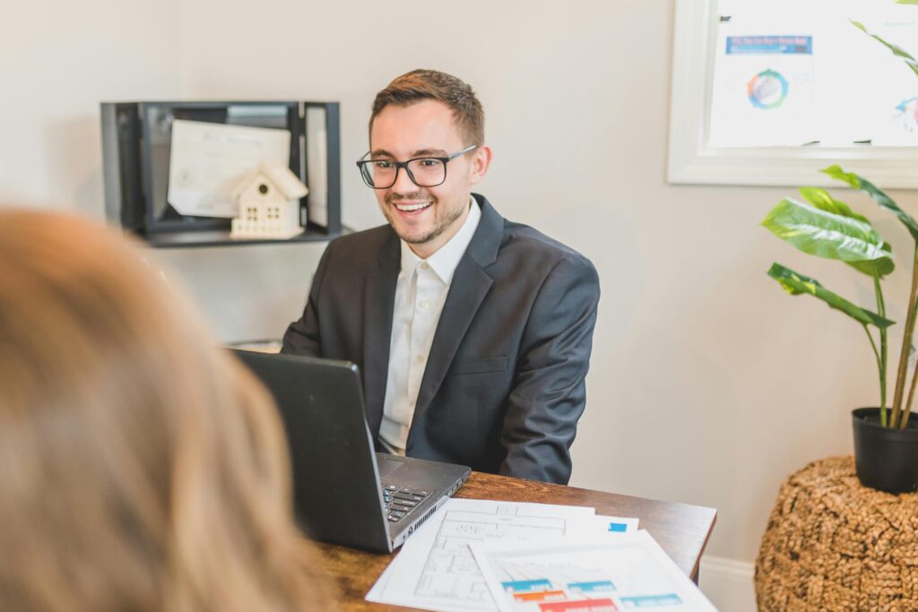 Business professional smiling while meeting with a client in an office setting, working on a laptop with documents on the desk.