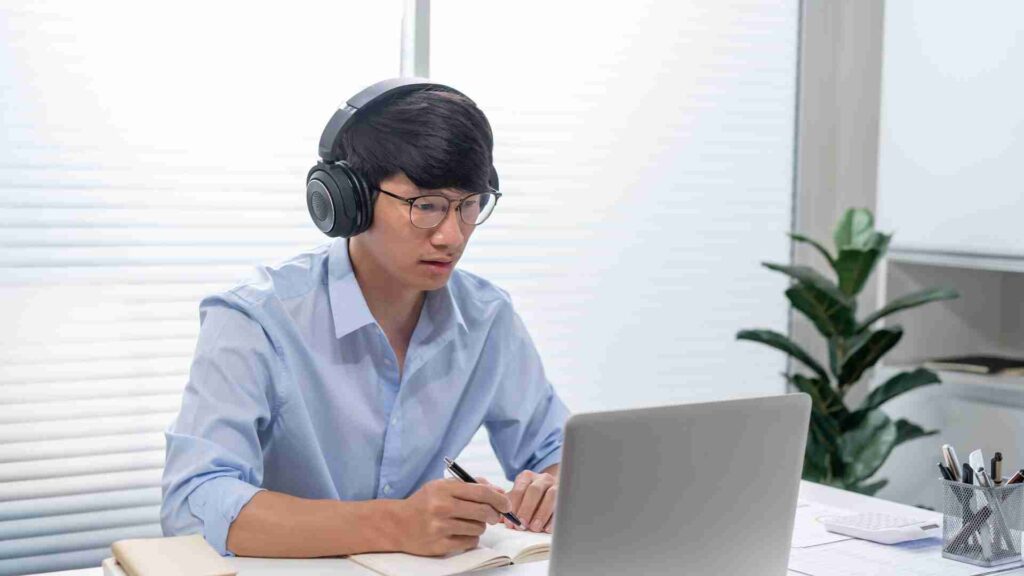 Person wearing headphones and glasses working on a laptop at a desk, taking notes in a notebook in a bright home office.
