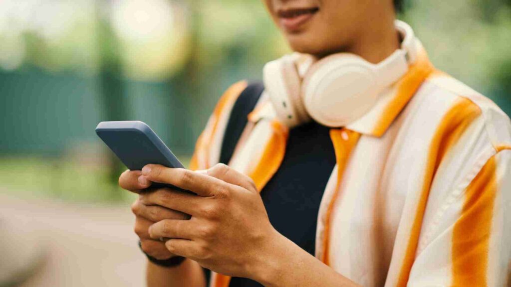 Person holding a smartphone outdoors while wearing headphones around their neck, checking their phone with a blurred green background.