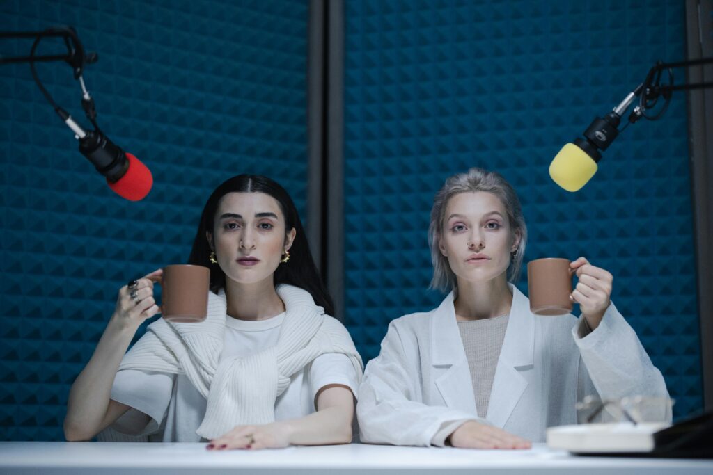 Two women recording a podcast in a soundproof studio, holding coffee mugs with microphones in front of them