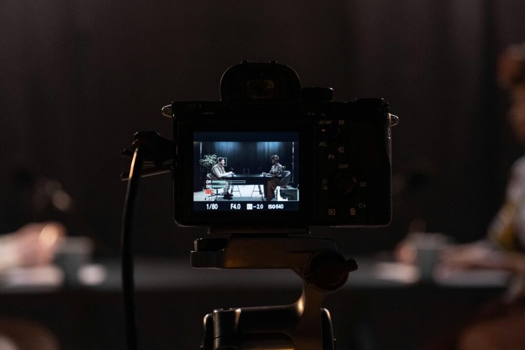 Alt text: Professional camera recording an interview setup with two people seated at a table under studio lighting, representing video content creation, podcast production, digital storytelling, and media content recording for online platforms.