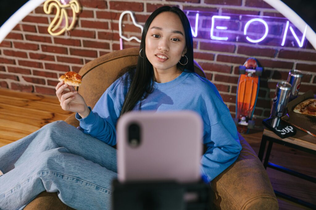 a girl sitting in front of ring light and phone, eating pizza and talking to followers