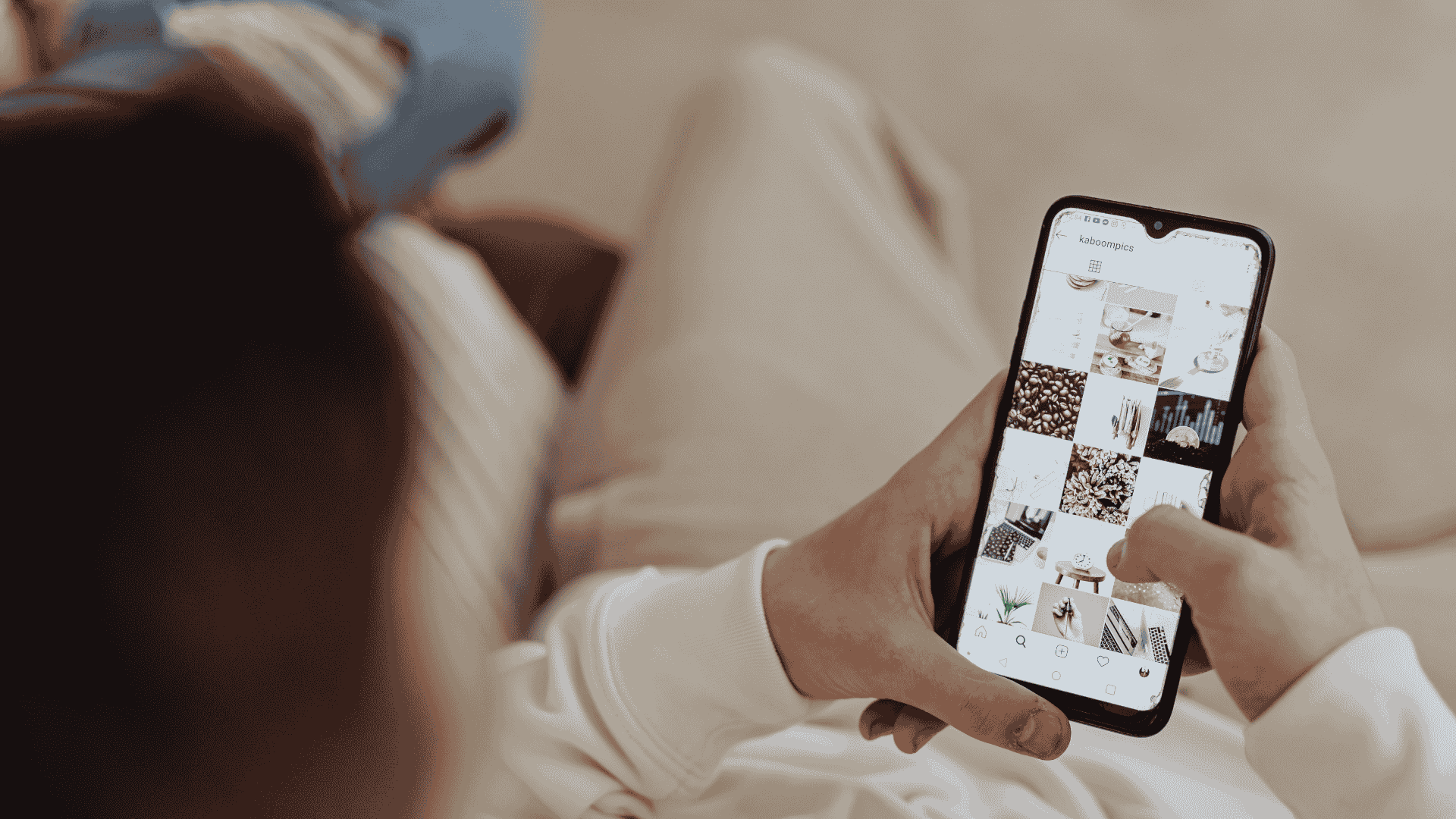 Person holding a smartphone and browsing an Instagram-style photo grid while sitting indoors in a relaxed setting.