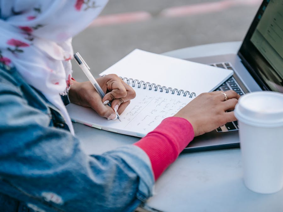 Crop anonymous Muslim woman student in casual clothes and headscarf sitting at table with cup of takeaway coffee and taking notes in notebook while browsing laptop in daytime