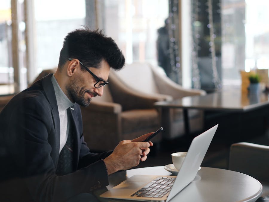 Professional man in café using smartphone with laptop open, smiling.