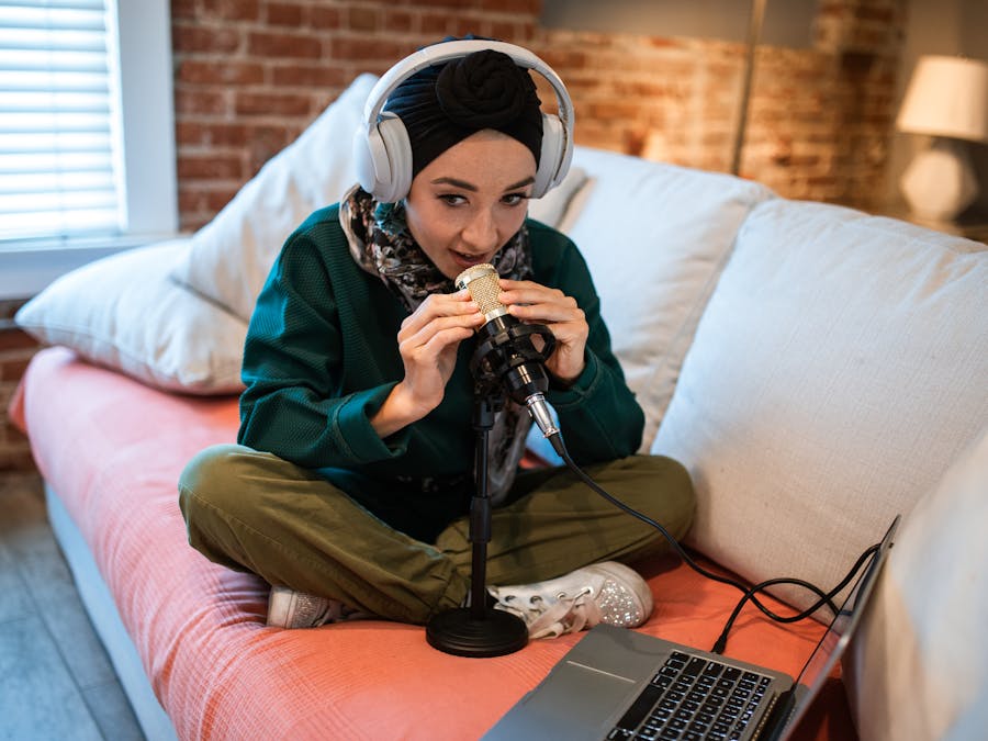 A young woman wearing headphones records a podcast at home using a microphone and laptop.