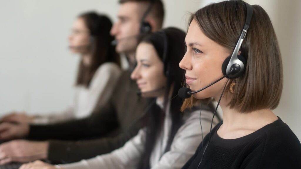 Side view of young coworkers in casual clothes sitting in light workplace with headphones with microphone while surfing on laptop