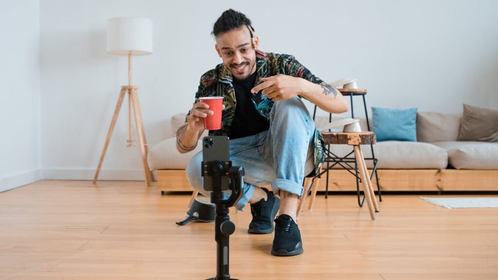 Man filming vlogs in a living room setting, holding a red cup and gesturing at smartphone on tripod.