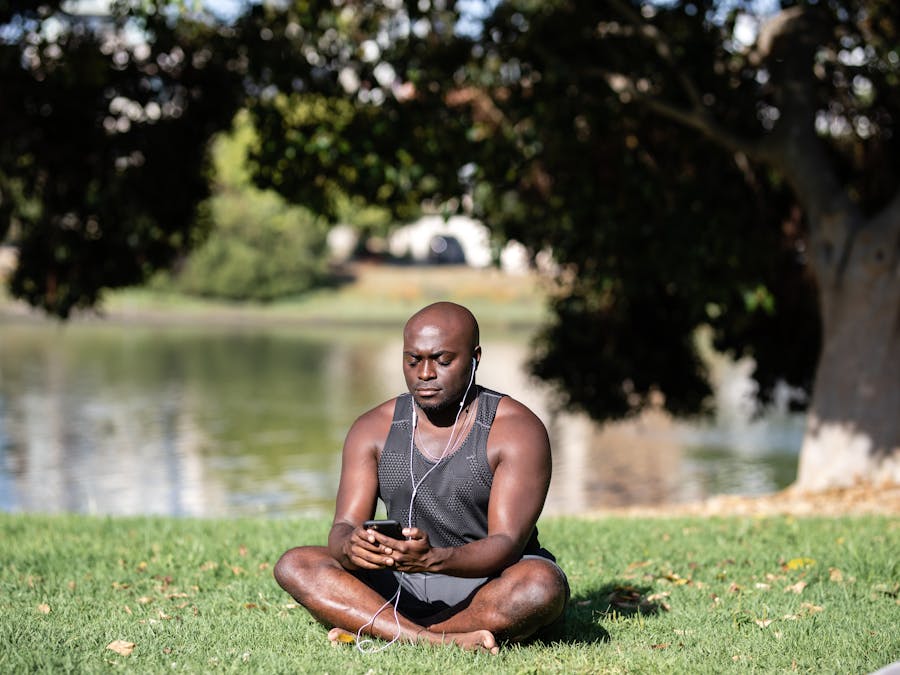 Adult man sitting cross-legged on grass, meditating with smartphone and earphones outdoors.