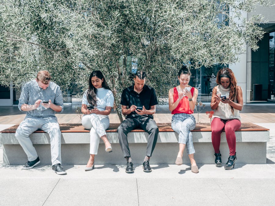 Group of diverse adults sitting outdoors using smartphones under a tree.