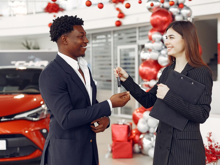 Side view of happy positive female dealer in formal classy wear passing car keys to excited African American customer in trendy suit in car showroom