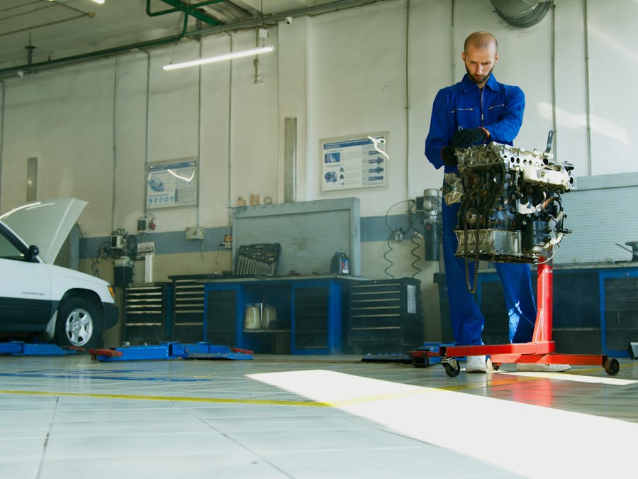 Mechanic lifting a car engine in a well-lit auto repair shop with vehicle in background.