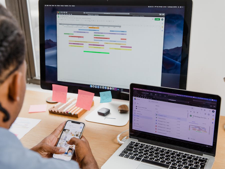 Businessman multitasks with smartphone, laptop, and monitor in office setting.