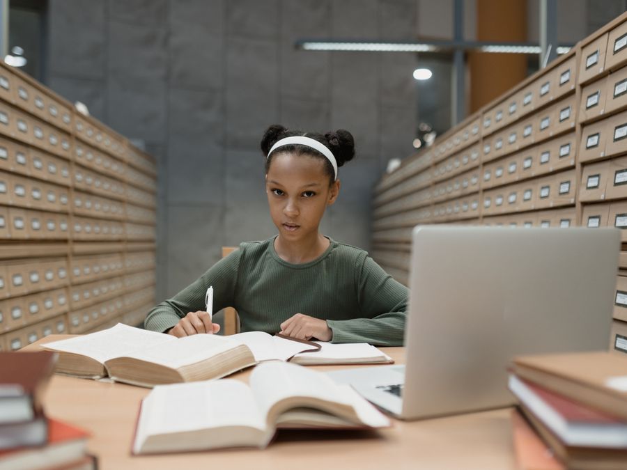 A focused young girl studying at a library desk with books and a laptop.