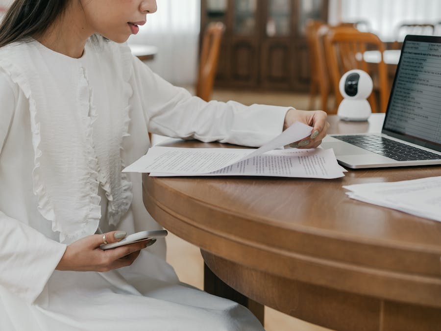 Woman in white dress studying at a wooden table with a laptop and papers indoors.