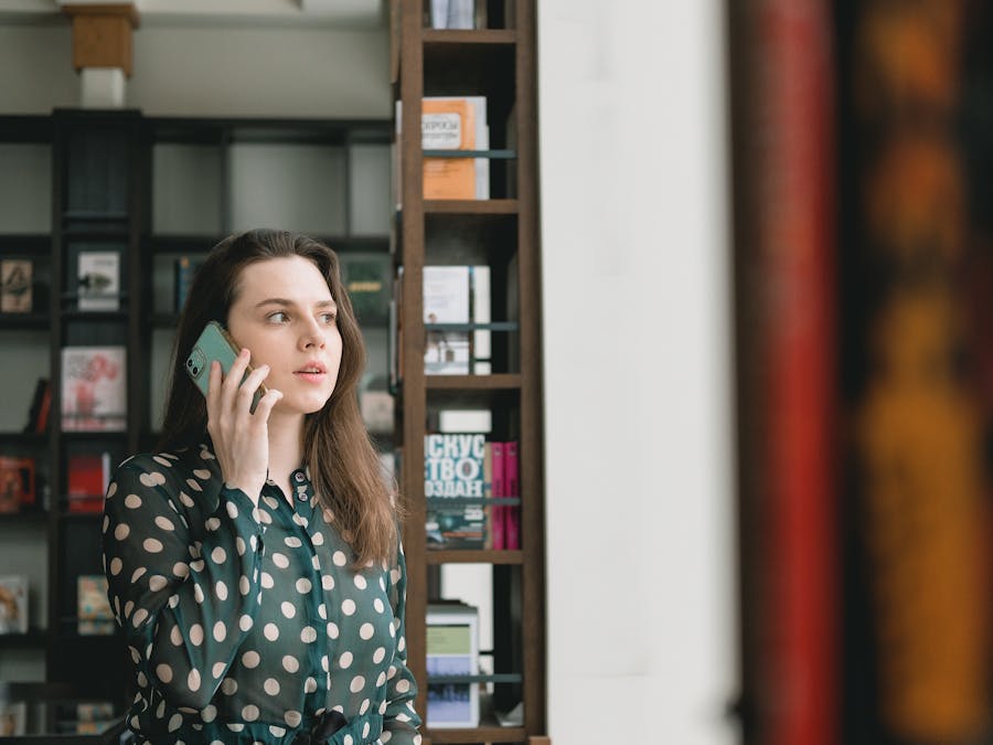 Elegant woman in polka dot dress talking on phone, standing in a modern library setting.