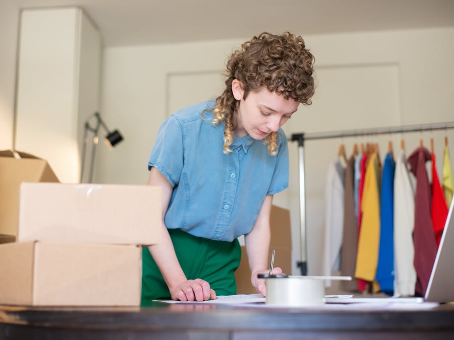 Woman packing online shop orders at home. Features boxes, clothing rack, and a laptop.