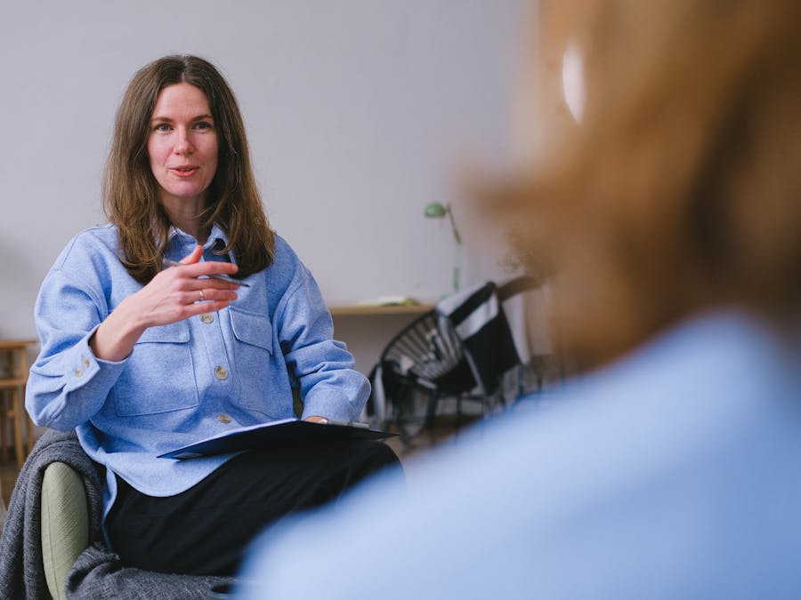 Woman in a blue jacket sitting indoors, engaged in conversation with another person.