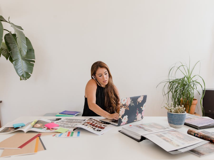 Woman working on a laptop while multitasking with calls, surrounded by creative materials and indoor plants in a home office.