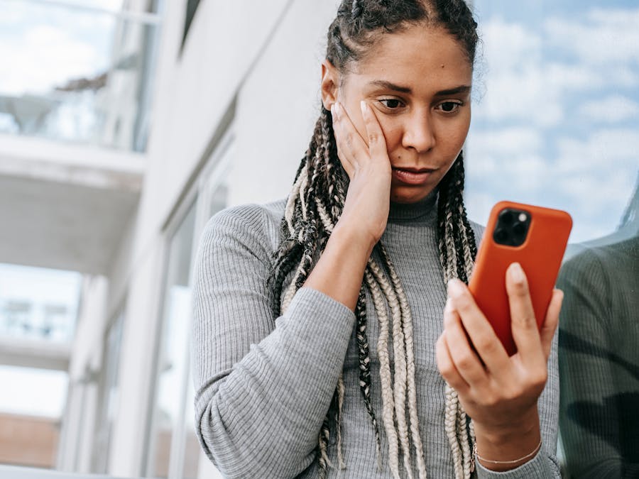 Crop serious African American female with dreadlocks touching cheek in contemplation and browsing mobile phone on street