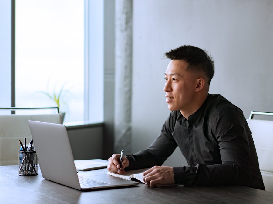 Asian male professional working at a desk in a modern office with a laptop and notepad.