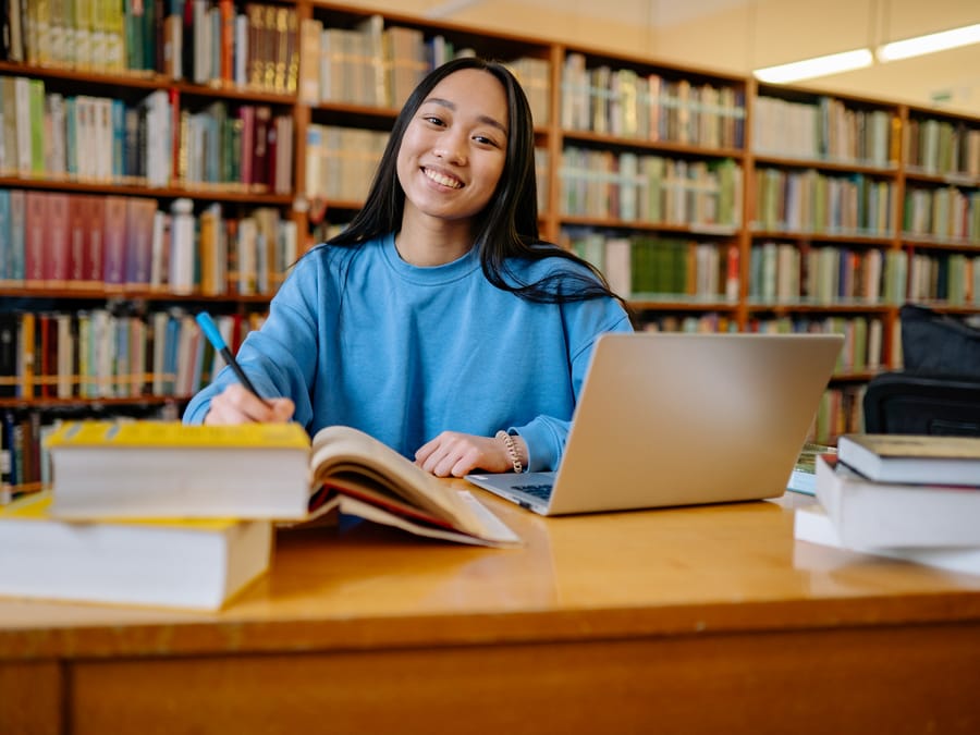 Smiling student studying in a library with books and laptop, showcasing education and focus.