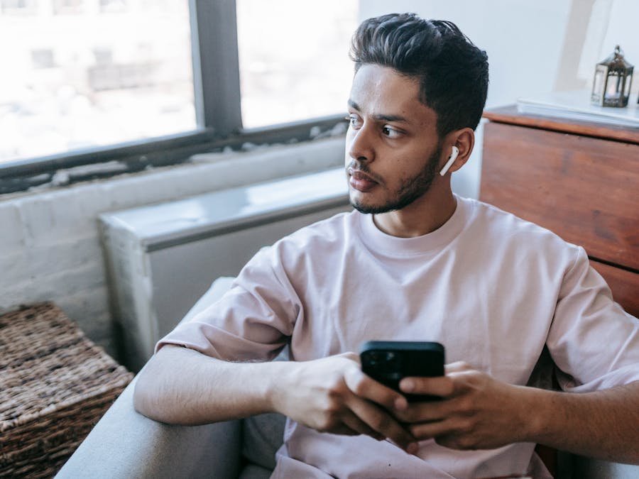 Focused bearded Indian male looking away and listening to music in wireless earphones while texting message on cellphone in armchair