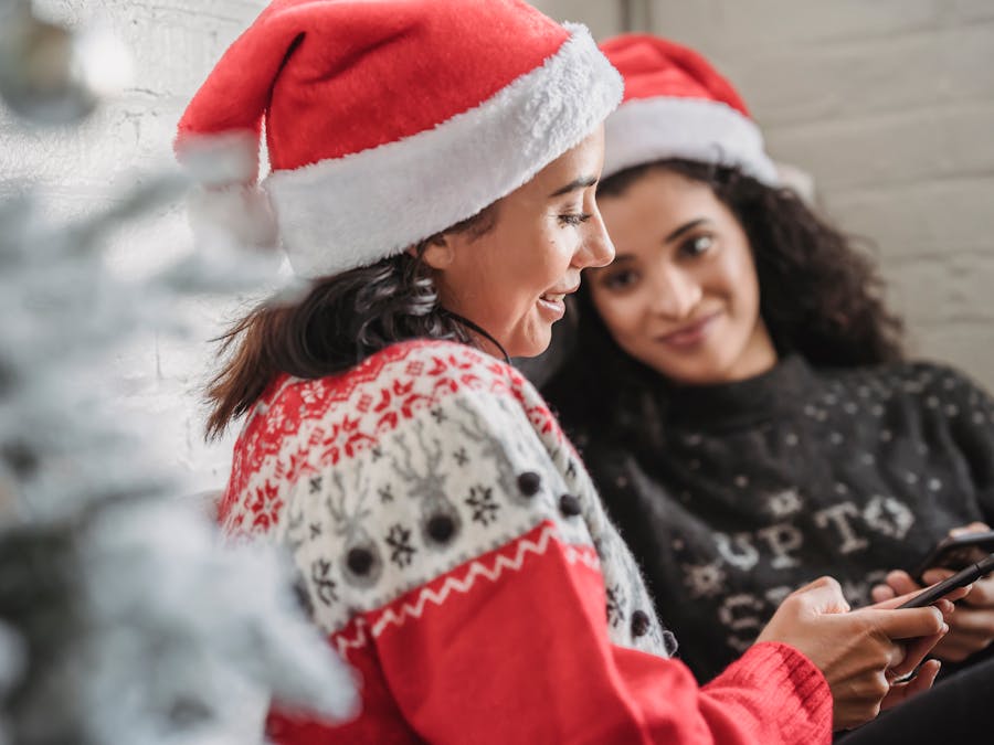 Side view of young smiling Hispanic female in Santa hat browsing mobile phone while celebrating Christmas holiday with female friend