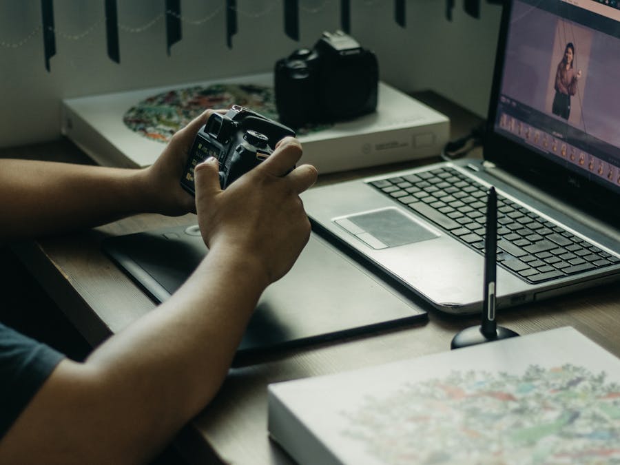 Photographer using a laptop and stylus for photo editing, holding a camera in an indoor setup.
