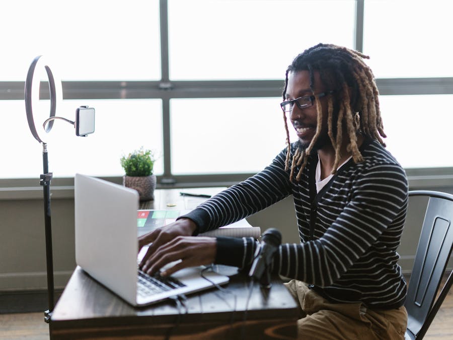 African American man with dreadlocks working on laptop indoors, using ring light and microphone.