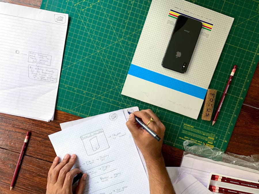 Designer drafting sketches on graph paper with smartphone and pencils on a wooden desk.