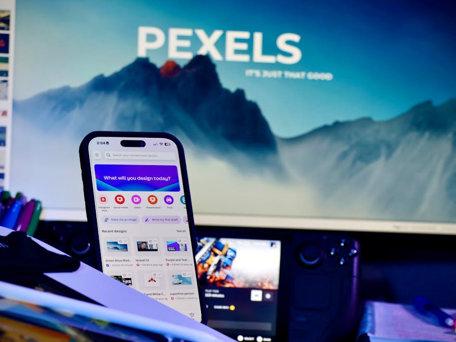 A creative workspace with a smartphone displaying design apps and a large monitor in the background.