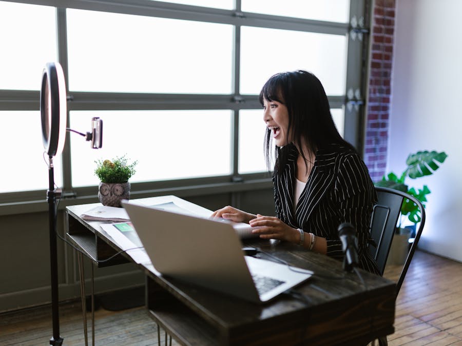 Asian woman in office setting recording content with laptop and ring light, showcasing elegant business setup.
