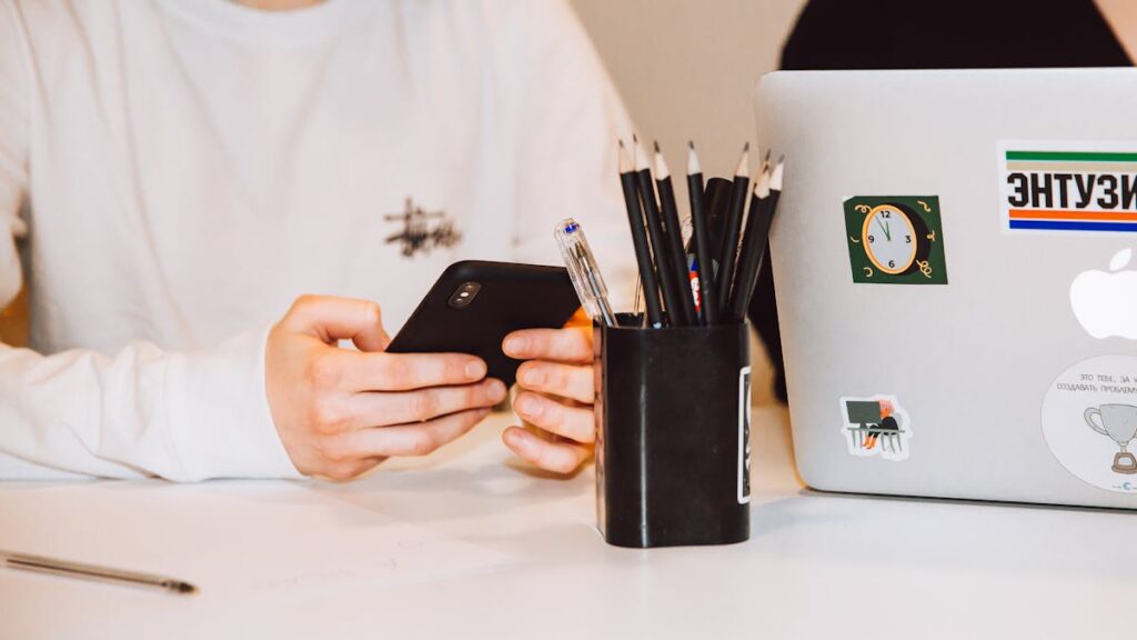 Young adult at a desk texting on smartphone, next to a laptop and stationery holder.