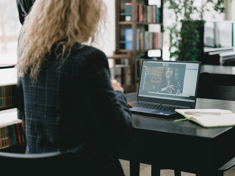 A woman sits at a laptop in a stylish library setting, engaging in a video call.