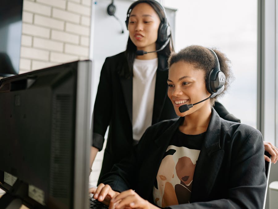 Two women with headsets engaged in customer service at a computer in a bright office.