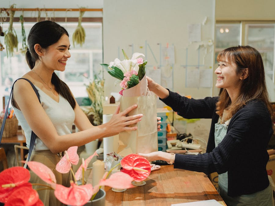 Side view of cheerful florist in apron giving paper bag with bouquets of flowers to smiling customer in floral shop in daytime
