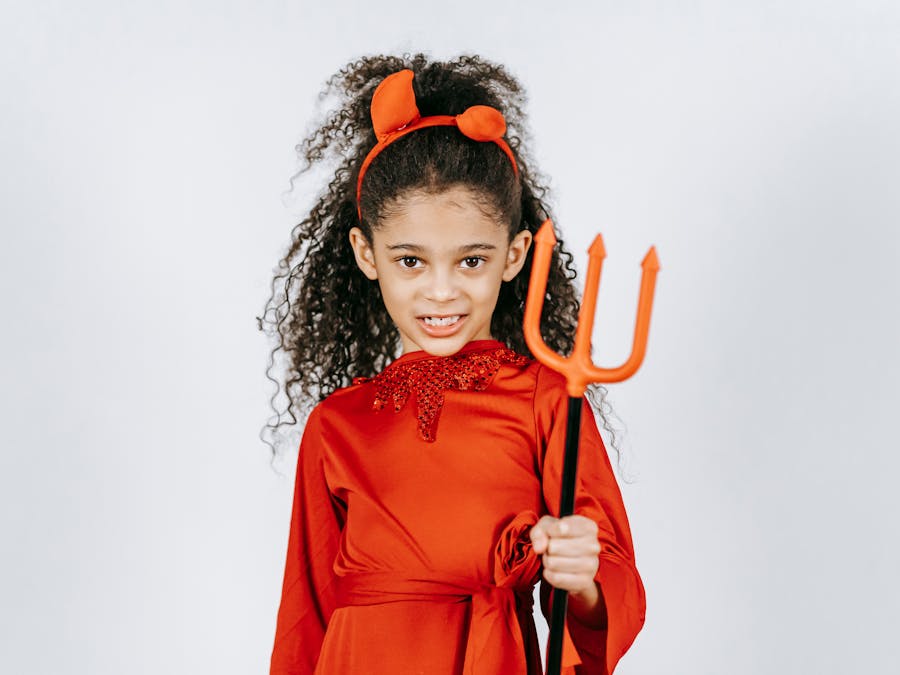Funny little African American girl in red costume and horns of devil holding trident and looking at camera against white background