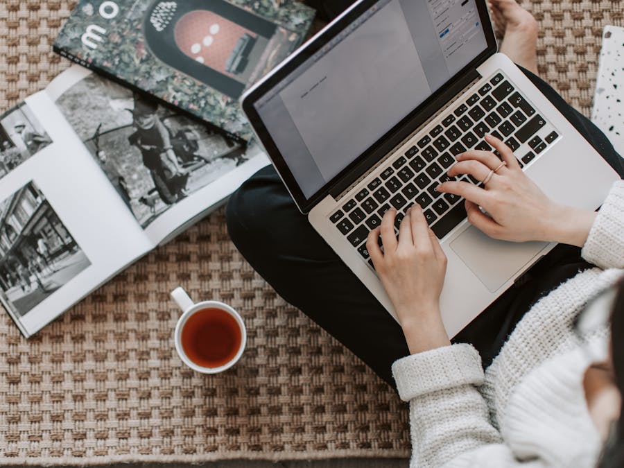 Top view of anonymous female freelancer in warm sweater drinking tea from white ceramic cup while sitting on floor carpet with netbook on legs near magazines and notebook and creating document for remote work project