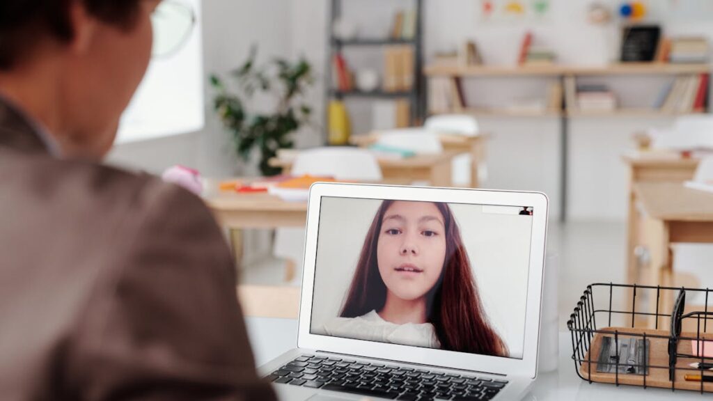 Teacher using laptop for virtual lesson with female student on screen in classroom setting.