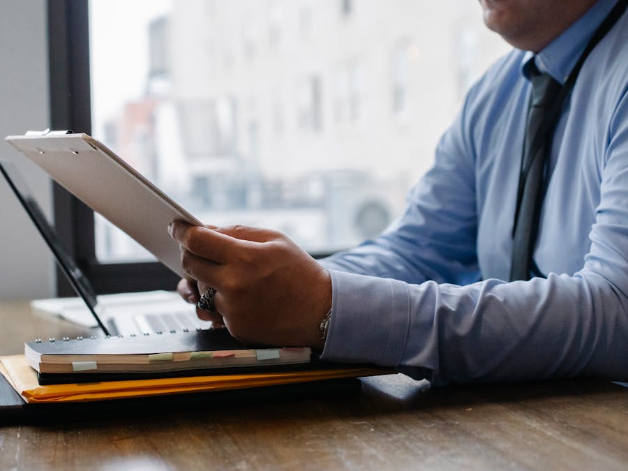 Crop unrecognizable male manager wearing blue shirt and black tie sitting at wooden desk and reading financial information on clipboard in office