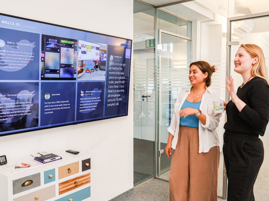 Two women in an office environment interact with a large digital display showcasing social media content.