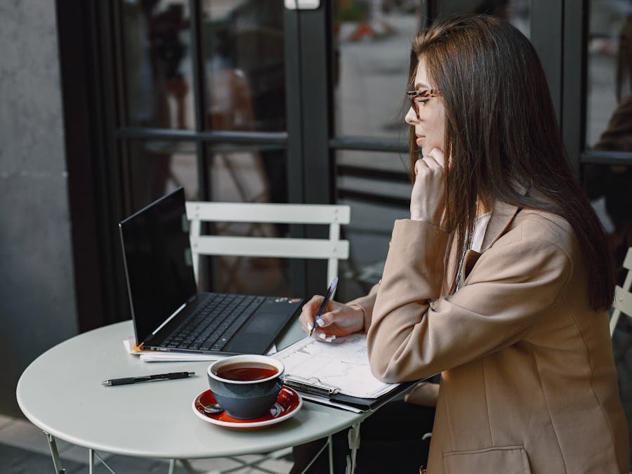 Woman writing in notebook at a coffee shop, using a laptop, with coffee.