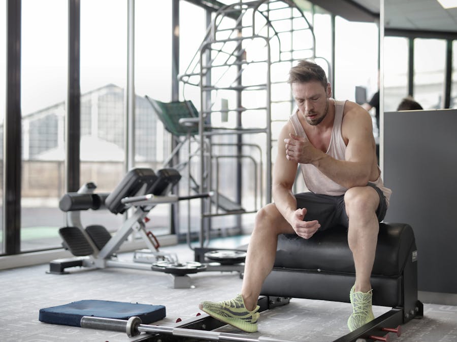 Tired young muscular male athlete in casual sportswear and sneakers sitting on bench and resting after intense weightlifting workout in modern fitness club