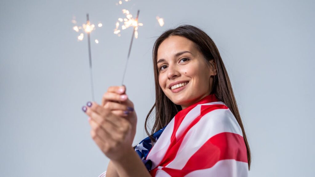 Young woman celebrating Independence Day with sparklers, wrapped in American flag.