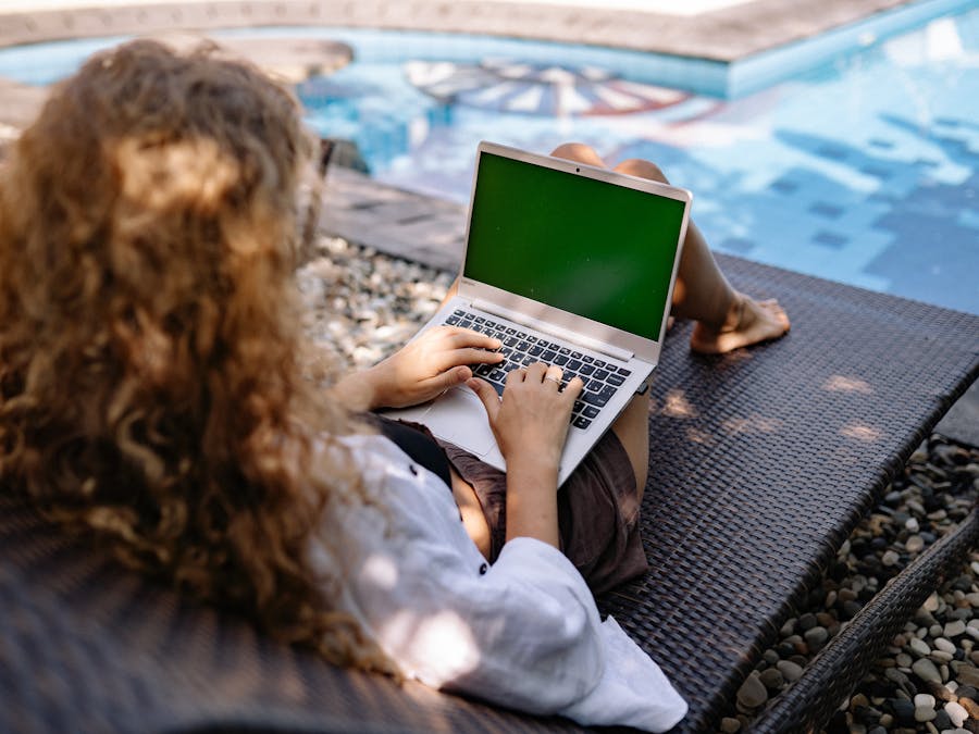 From above back view of anonymous female freelancer typing on portable computer while lying on sunbed near swimming pool with transparent water in sunlight