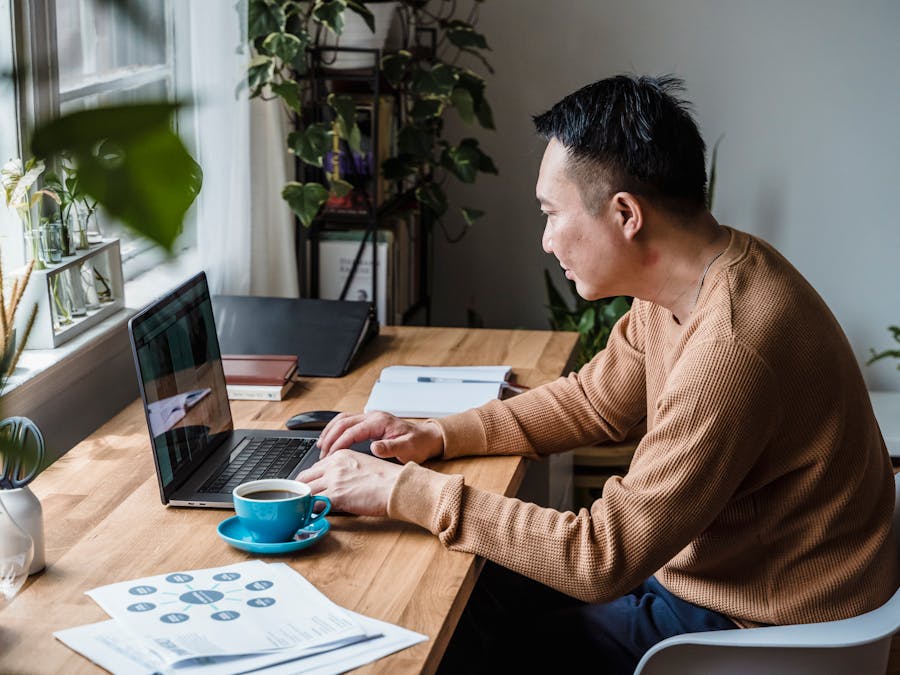 Man in brown sweater working on a laptop with coffee on a wooden desk in a home office setting.