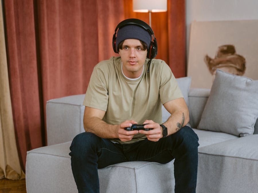 A young man wearing a beanie and wireless headphones, playing video games on a couch.