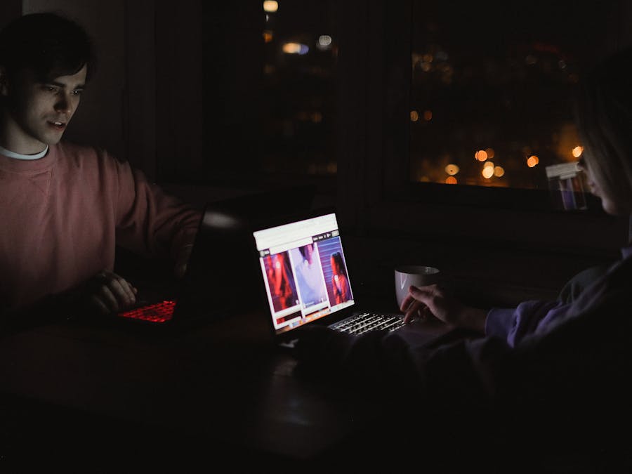 A man and woman using laptops at night, illuminated by screens in a dark, cozy room.