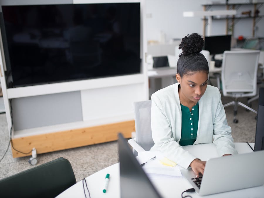 Focused woman using a laptop in a modern office setting, wearing a white blazer, working diligently.